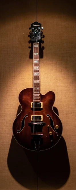 A brown Ibanez semi-hollow electric guitar mounted on a padded wall under warm lighting. A brown Ibanez semi-hollow electric guitar mounted on a padded wall under warm lighting.